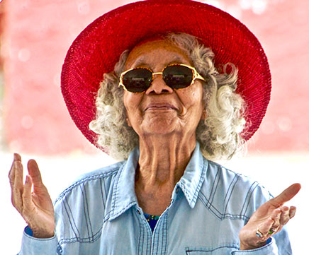 Uma senhora idosa muito estilosa, usa chapéu de abas grandes vermelho, óculos de sol e dá um sorriso discreto