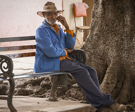 Senhor idoso está sentando no banco da praça. Ele usa chapéu e bota