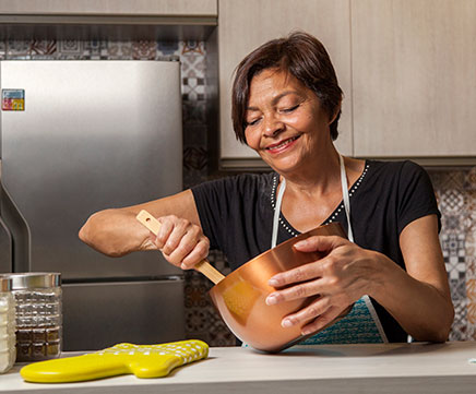 Uma senhora está em sua cozinha, mexendo com uma colher de pau em uma panela