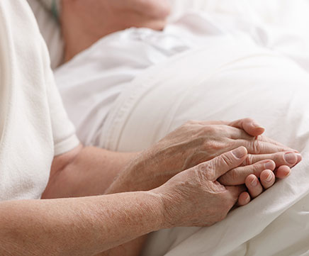 Um senhor idoso está de cama, enquanto uma senhora segura sua mão com carinho