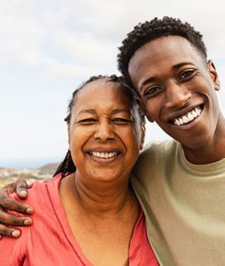 Uma senhora idosa recebe o abraço apertado de um jovem. Os dois sorriem animados