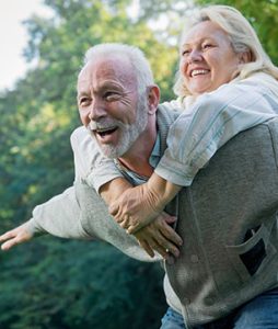 Um casal de idosos estão brincando animados de aviãozinho. Ele imita o avião e ela está de cavalinho nas costas dele