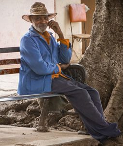 Senhor idoso está sentando no banco da praça. Ele usa chapéu e bota